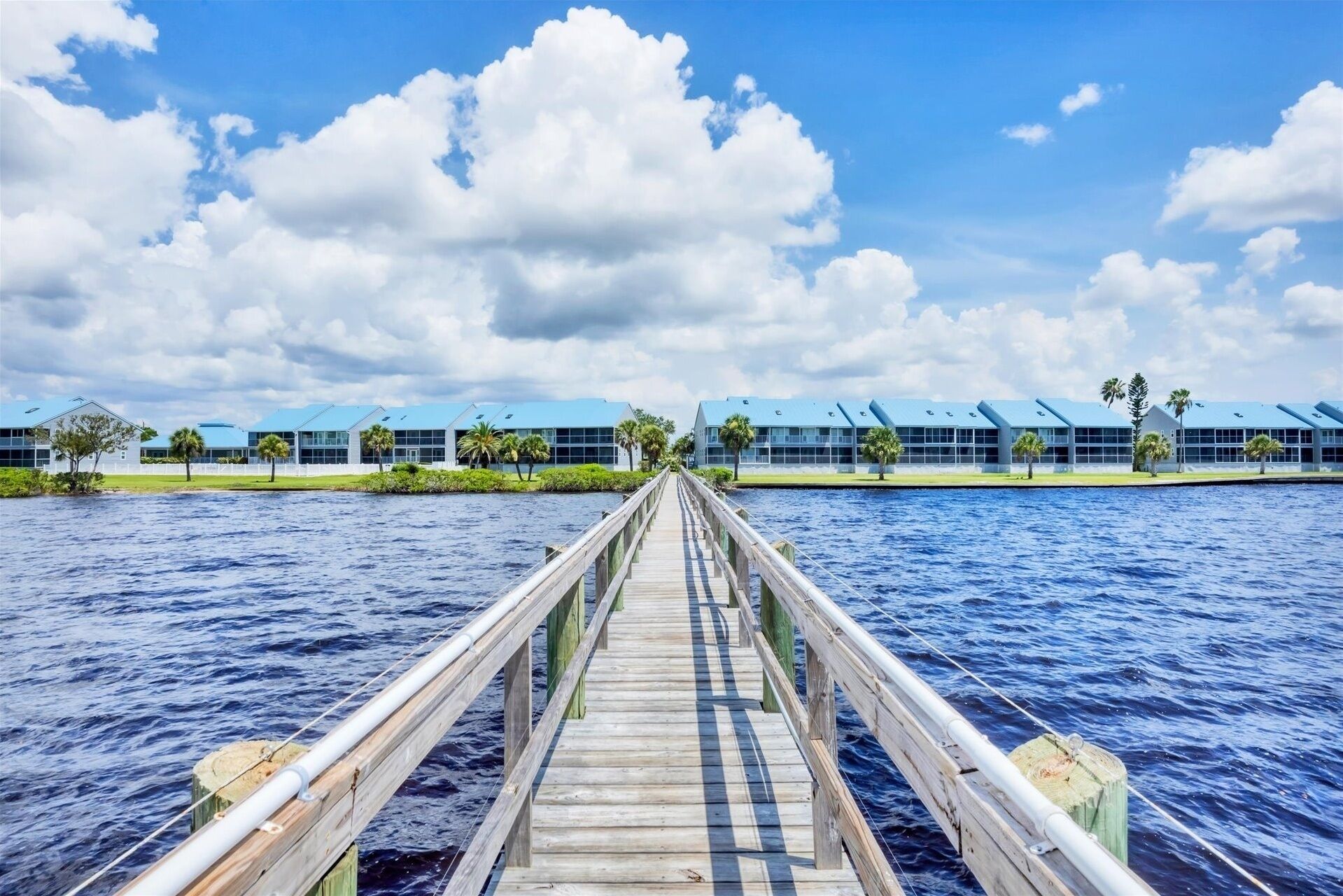 A wooden pier leading to a row of modern buildings with blue roofs, overlooking a body of water.