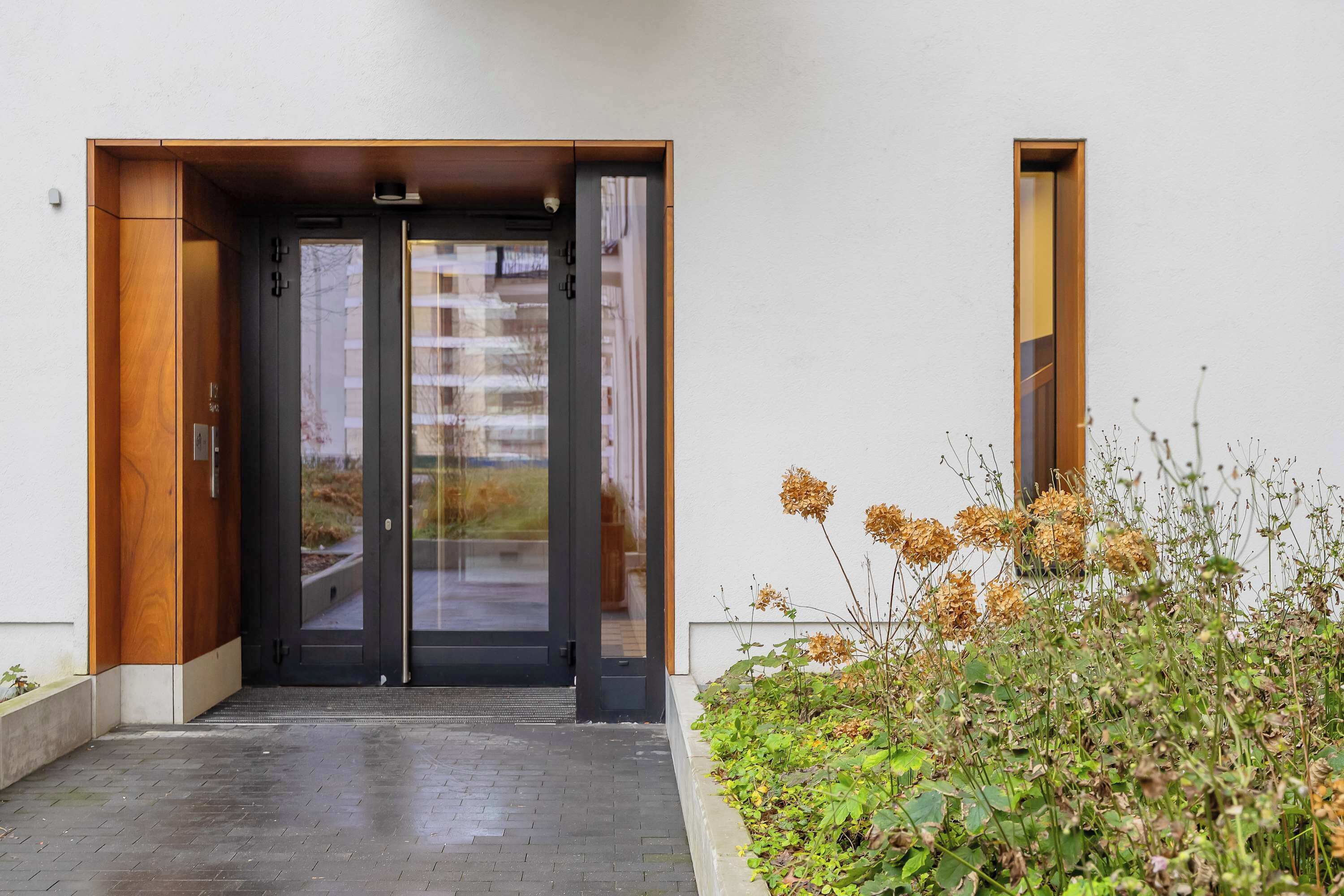 A modern entrance with a glass door, a wooden frame, and a small garden area with plants and dried flowers.