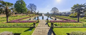 Ein formaler Garten mit einem zentralen Brunnen, symmetrischen Blumenbeeten und blauem Himmel.