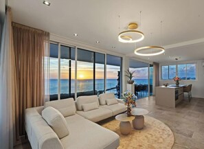 A modern living room with a large sofa, round coffee table, and a view of the ocean at sunset. - Penthouse • Oceanfront 2BR Luxury • ONE Mambo 36 (Willemstad)