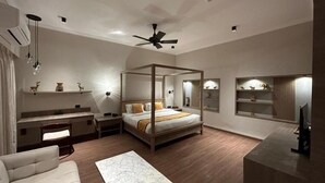 A modern bedroom with a four-poster bed, a sofa, a marble coffee table, a TV, and a ceiling fan. - Narwar Lakefront Resort (Shivpuri)