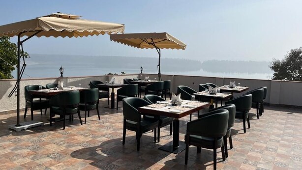An outdoor dining area with tables and chairs, covered by umbrellas, overlooking a body of water. - Narwar Lakefront Resort (Shivpuri)