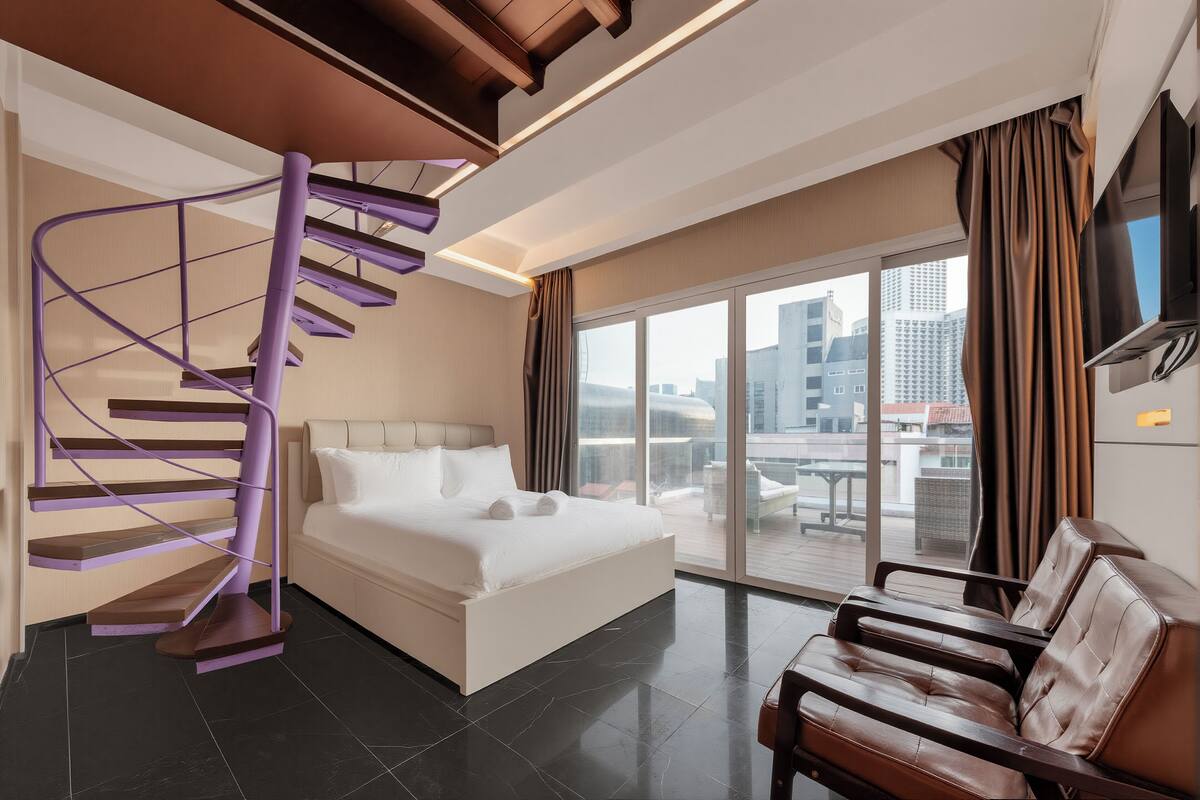Ein modernes Hotelzimmer mit einem weißen Bett, einer violetten Wendeltreppe, einem Ledersofa und einer großen Schiebeglastür, die auf einen Außenbalkon führt.
