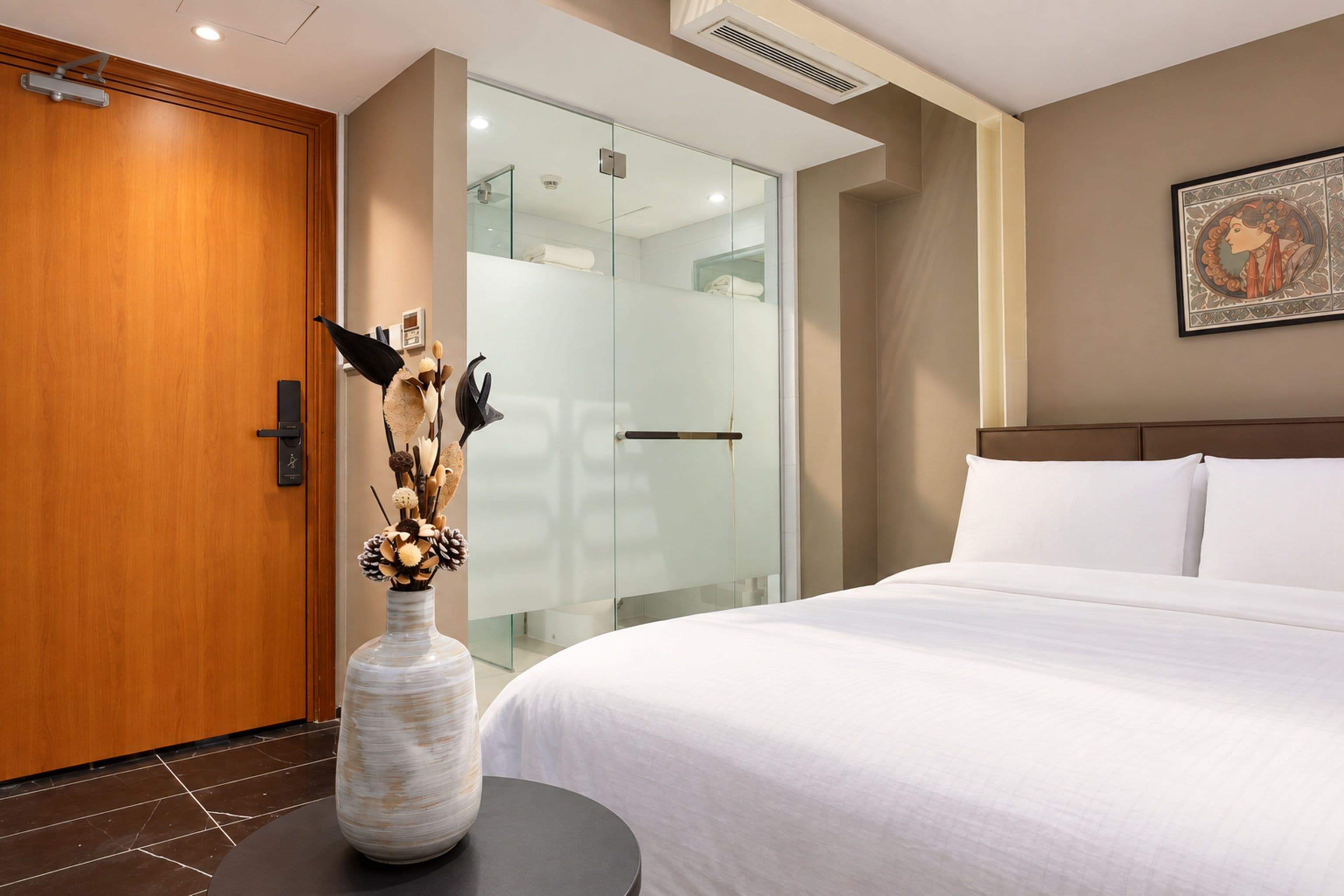 A hotel room with a wooden door, a glass shower enclosure, a bed with white bedding, and a vase with dried flowers.