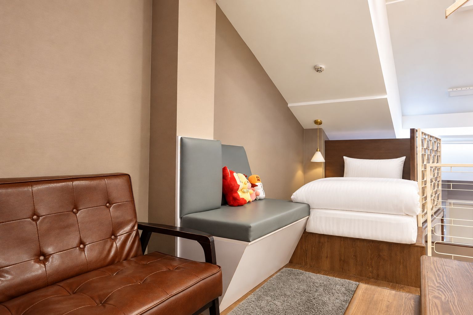 A hotel room with a brown leather sofa, a grey cushioned bench, a bed with white linens, and a small table with plush toys.