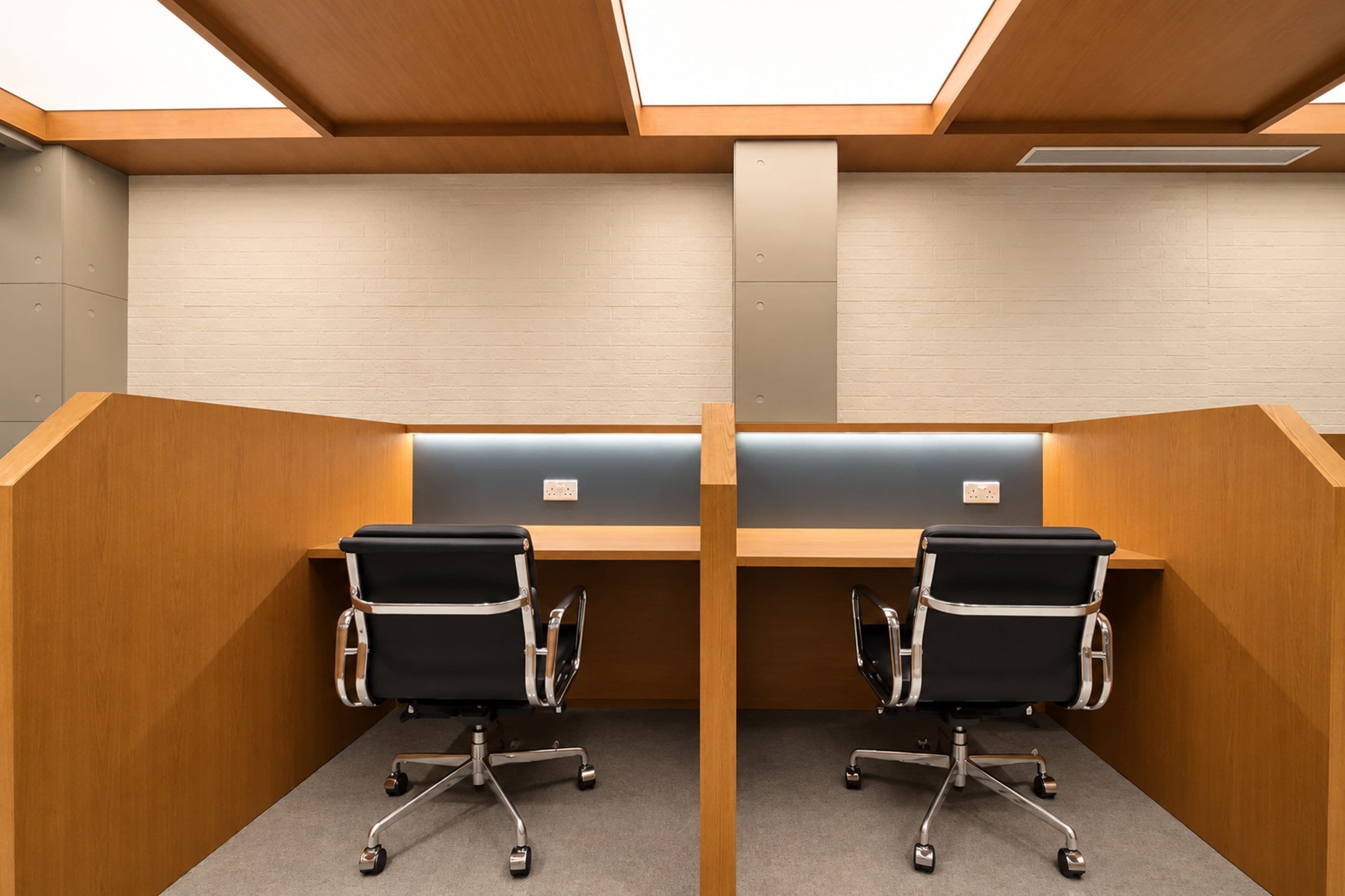 A modern office cubicle with two black office chairs, a wooden desk, and a white brick wall.