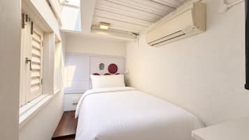 A compact hotel room with a bed, a small desk, an air conditioning unit, and a window with shutters.