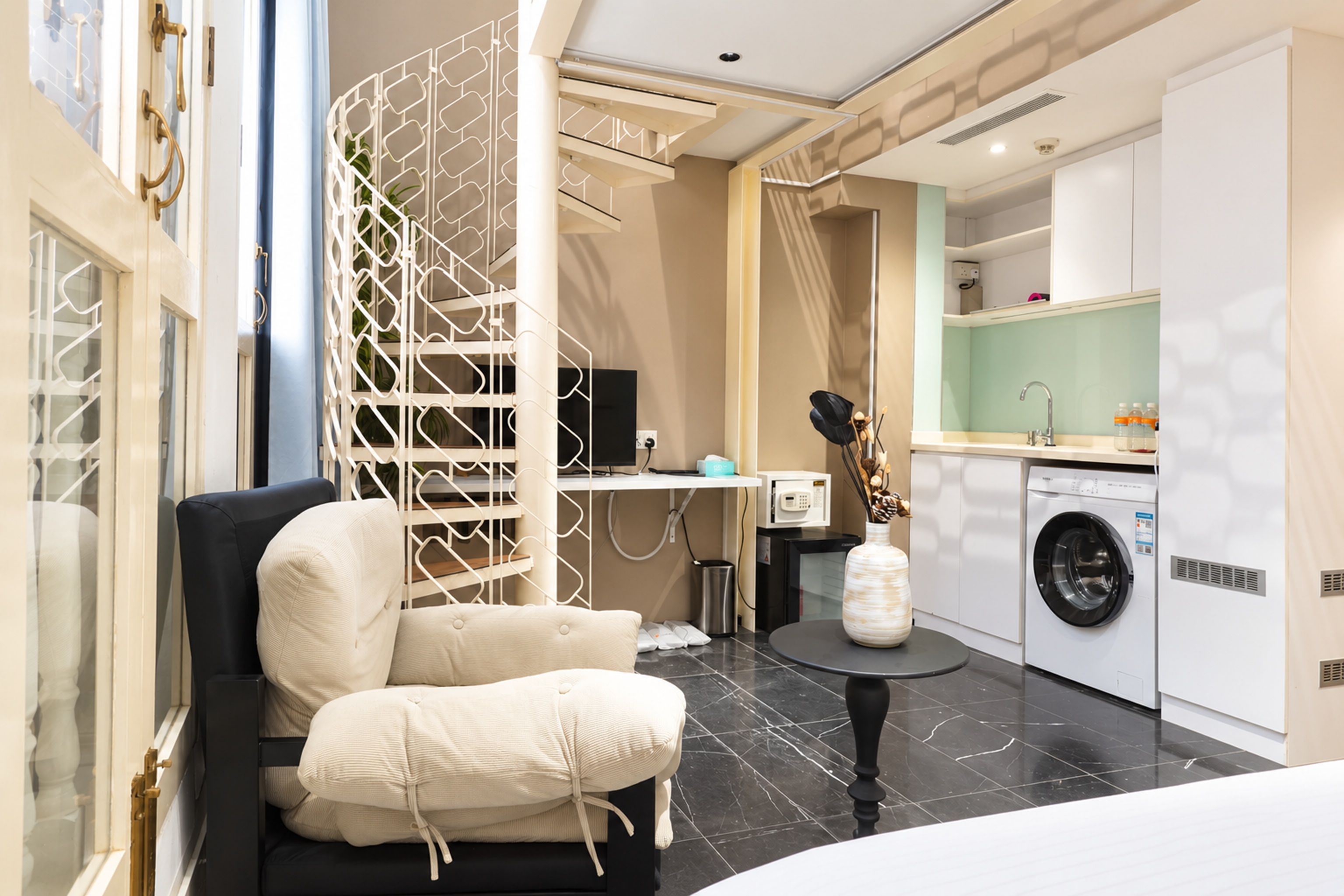 Een moderne hotelkamer met een zwarte leren bank, een witte decoratieve scheidingswand, een wasmachine en een klein rond tafeltje met een vaas.