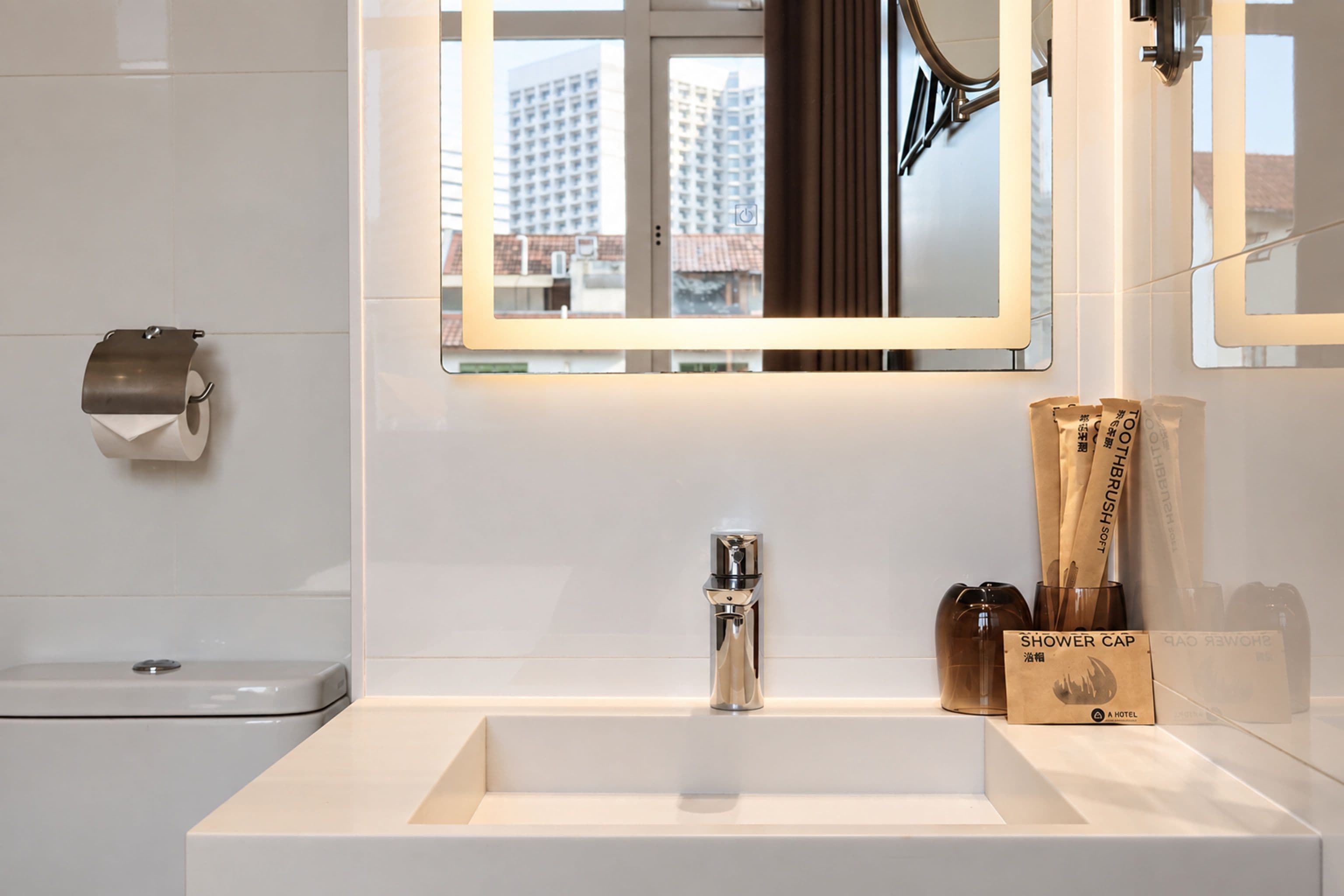 Un bagno moderno con un lavandino bianco, un rubinetto cromato e uno specchio sopra. Sul mobile ci sono articoli da toilette e si intravede una vista sugli edifici esterni.