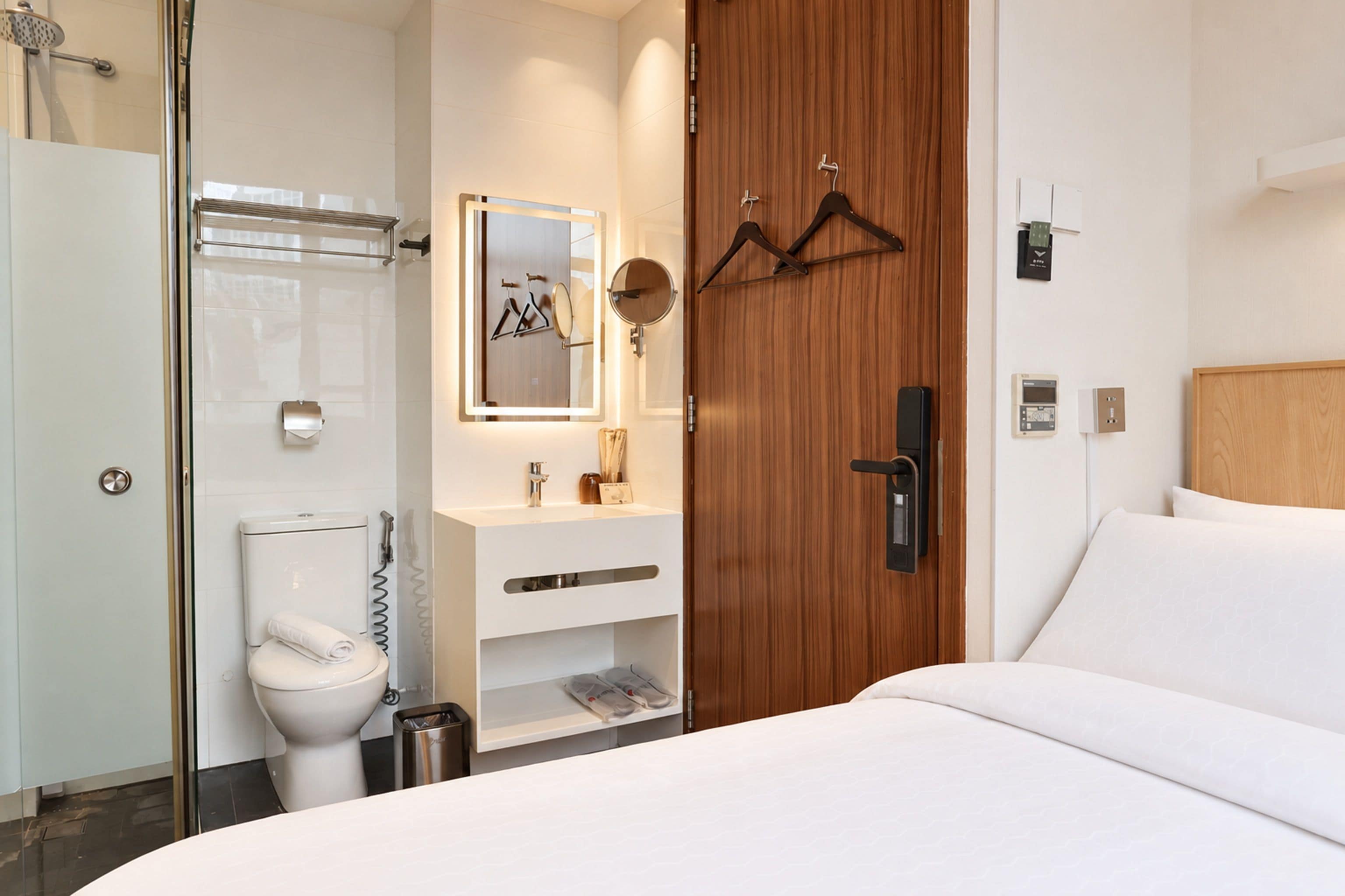 A modern hotel room with a bed, a wooden wardrobe, a bathroom with a toilet, a sink, and a shower.
