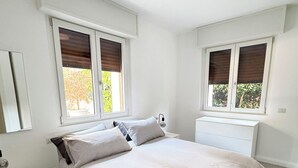 A bedroom with a large bed, two windows with blinds, a bedside table, and a nightstand. - Casa Mina (Opera)