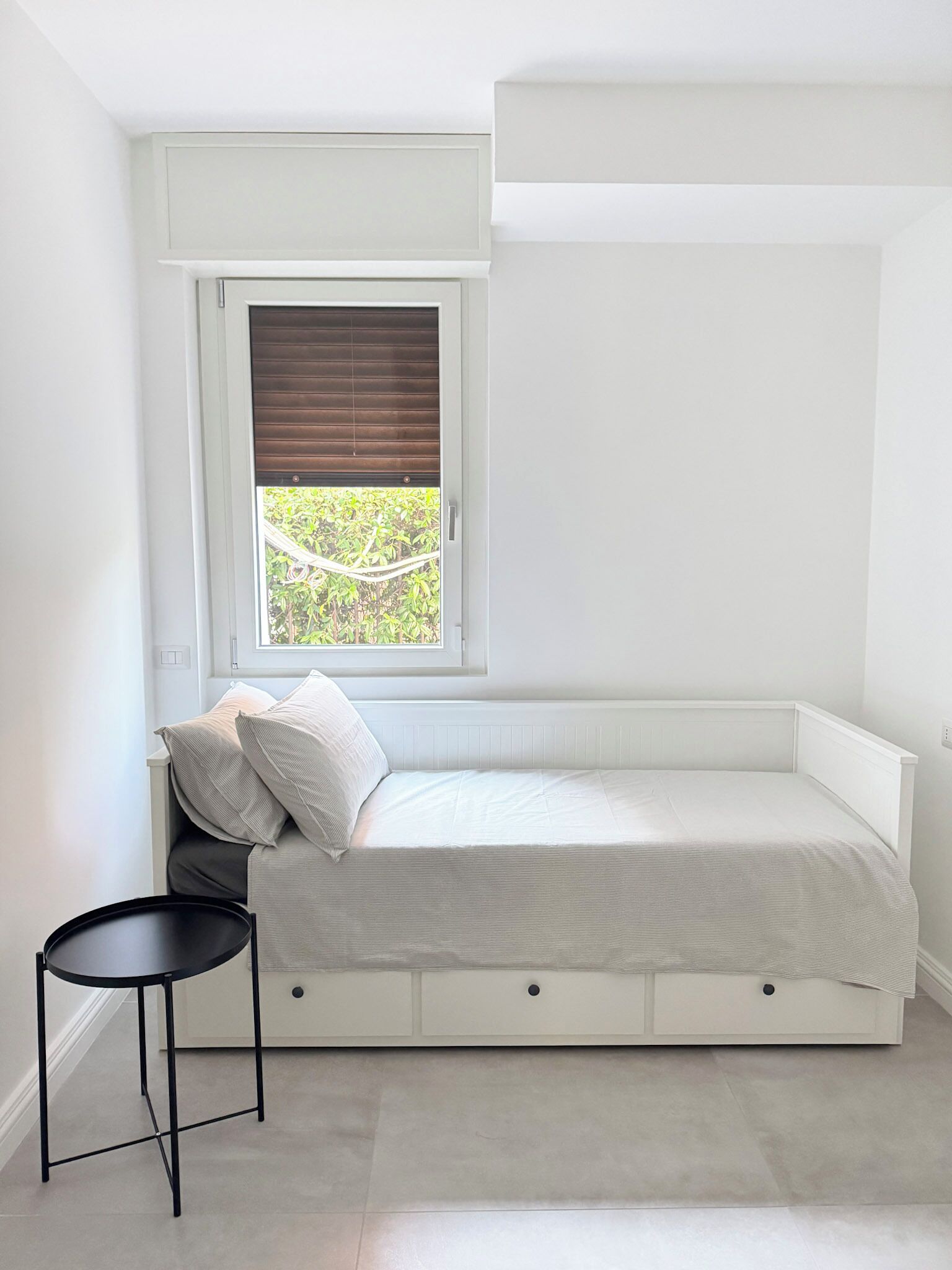 Ein kleines Schlafzimmer mit weißen Wänden, einem Einzelbett, einem Fenster mit Jalousien und einem kleinen schwarzen Tisch.