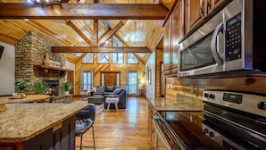A spacious kitchen with granite countertops, a stone fireplace, and a large window overlooking the living area. - Treetop Hideaway, Shuffleboard, Hot Tub, Bunks, Games! Free Firewood (Broken Bow)