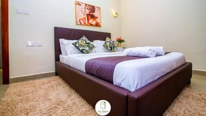 Un lit bien fait, avec une tête de lit violette, une literie blanche et des coussins à motifs. On peut voir des appliques murales et un tableau encadré au-dessus du lit.