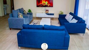 Un salon comprenant un grand canapé bleu, une table basse blanche et un téléviseur à écran plat fixé au mur.