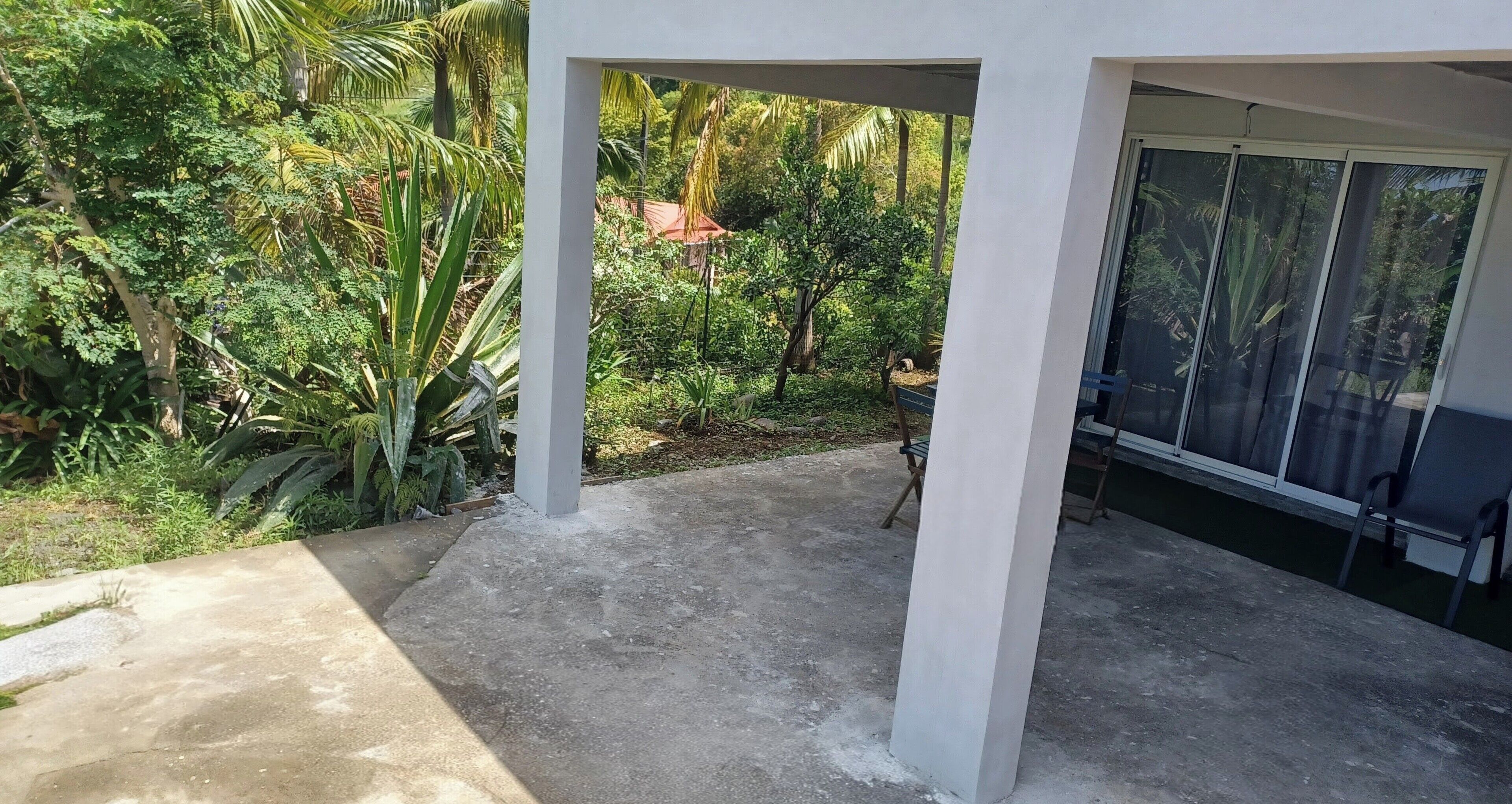A covered outdoor area with concrete flooring, surrounded by lush greenery and a view of a house in the background.