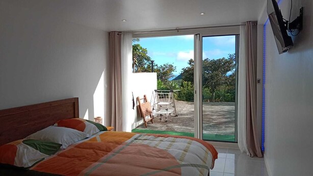 Une chambre à coucher avec un lit, une porte-fenêtre coulissante donnant sur une terrasse équipée d’une table et de chaises, et un projecteur fixé au mur.