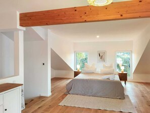 Un dormitorio con techo inclinado, una cama con ropa de cama blanca, dos mesitas de noche con lámparas y un ventanal con vistas a la vegetación.