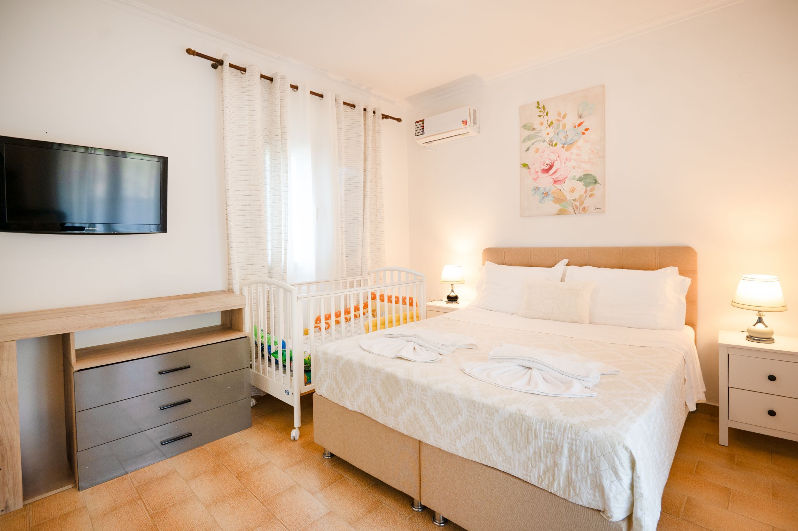 A bedroom with a bed, bedside tables, a TV, a crib, and a wall-mounted air conditioner.