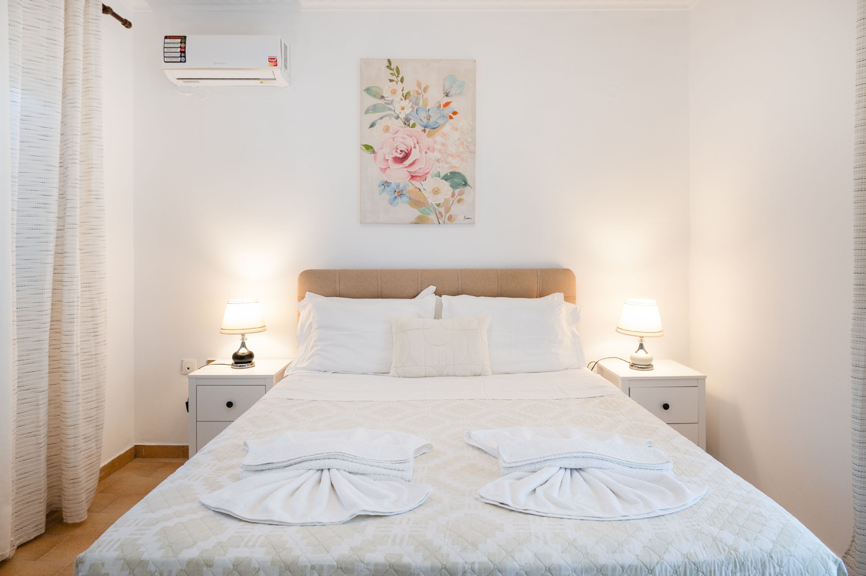 A neatly made bed with two towels, a floral artwork on the wall, and a window with an air conditioning unit.