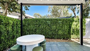 A covered outdoor seating area with a round table and stools, surrounded by a neatly trimmed hedge. - Family-Friendly Pool & Patio Retreat, Walk to Beach & Dining (Trinity Beach)