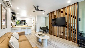 A modern living room with a sofa, a coffee table, and a TV. There is a kitchen area with a bar and stools. - Family-Friendly Pool & Patio Retreat, Walk to Beach & Dining (Trinity Beach)