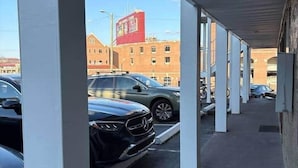 Ett överbyggt parkeringsområde med vita pelare, en svart bil parkerad och en röd byggnad i bakgrunden.