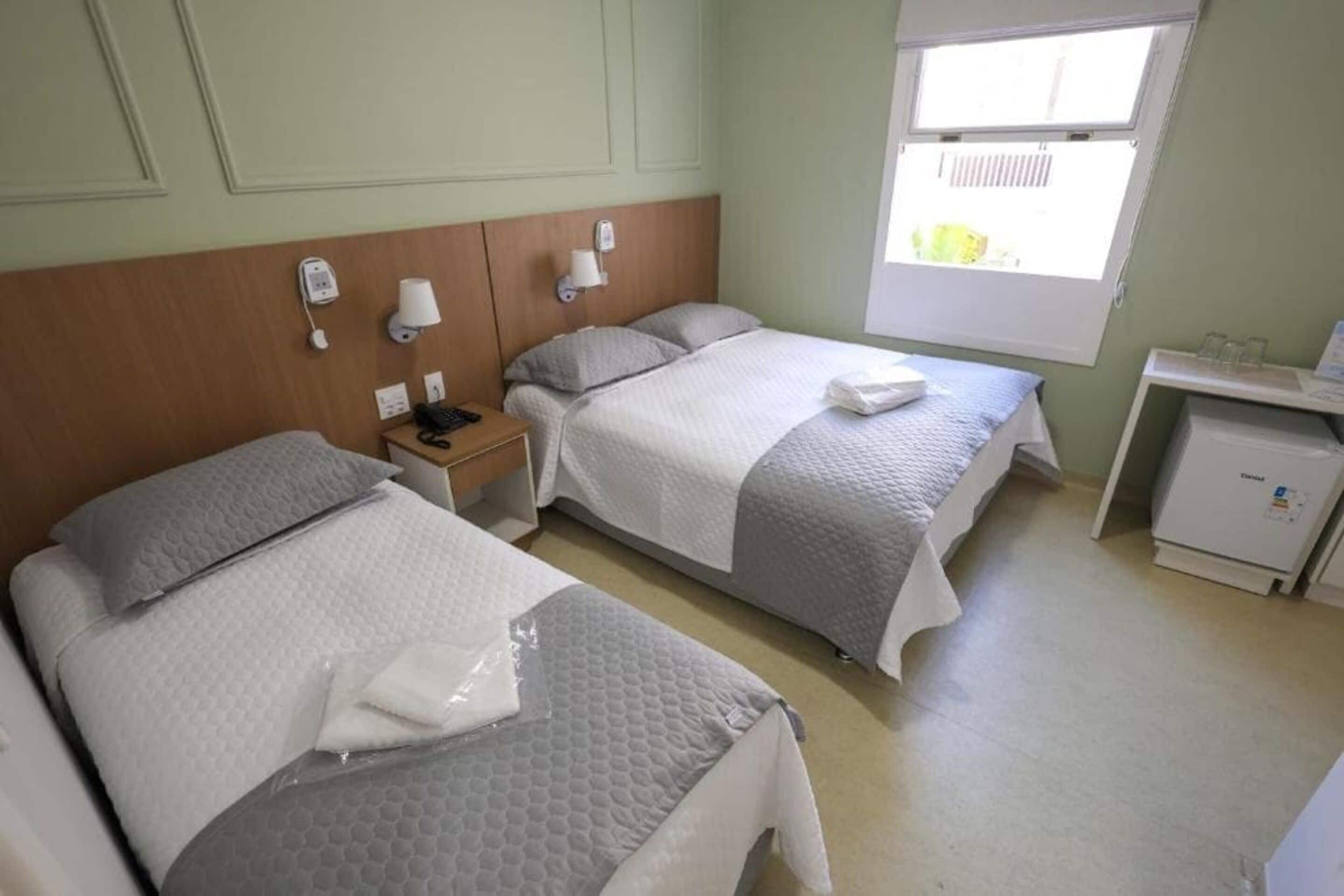 A hotel room with two beds, a small desk, a refrigerator, and a window with a view.