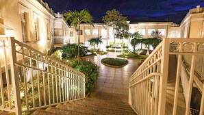 A well-lit courtyard with a central fountain, surrounded by buildings with balconies and a staircase leading to an upper level.