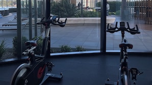 Un gimnasio con bicicletas estáticas y vistas a un paisaje urbano a través de amplios ventanales.