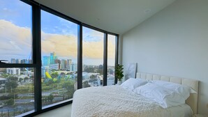 Un dormitorio con un ventanal grande, cama con ropa blanca, planta en maceta y vista a la ciudad.