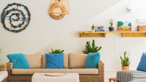 Ein gemütliches Wohnzimmer mit einer Couch, einem Hocker, Sesseln, einem gewebten Couchtisch, einem blauen Teppich, einer Wandlampe und Regalen mit Pflanzen und Dekoration.