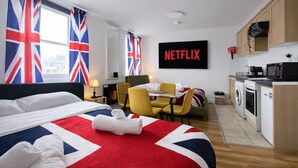 Ein Hotelzimmer mit Bett, kleinem Esstisch, Stühlen, einer Küchenzeile mit Geräten und einem an der Wand befestigten Fernseher.