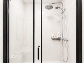 Doccia con soffione a mano e rubinetto a parete, vaso e ripiano con articoli da toilette.