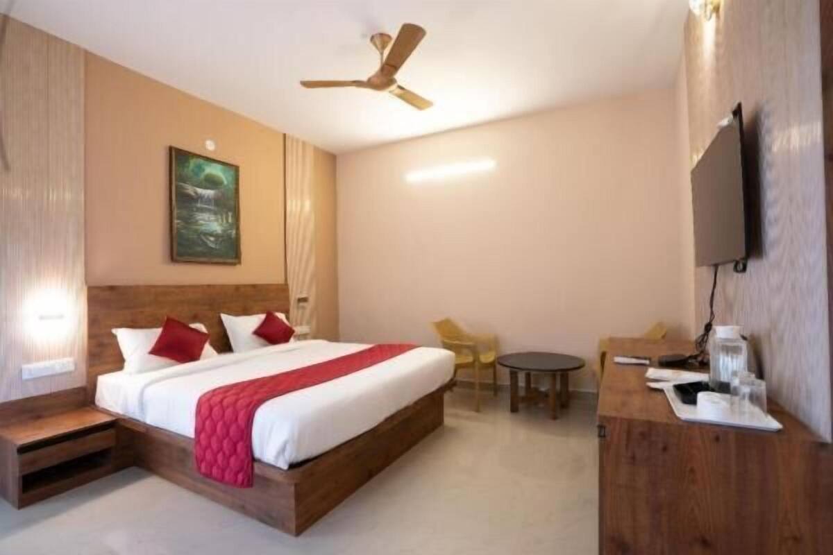 Habitación de hotel con una cama de madera, ventilador de techo, un cuadro, un escritorio con silla y televisor montado en la pared.