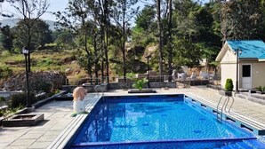 A swimming pool with a tiled deck, surrounded by trees and a small building with a blue roof. - MSB Silver Spring Resort Private Limited (Yelagiri)