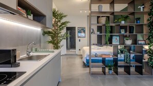 A modern kitchen with a built-in stove, a sink, and a countertop. There is a bed with a blue headboard in the background. - The Next Chapter Nilie Hospitality MGMT (Thessaloniki)