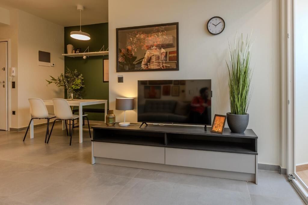 Een moderne woonkamer met een flatscreen televisie op een zwarte standaard, een eethoek met een tafel en stoelen, en een groene accentmuur.