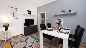 Un espace repas moderne avec une table blanche, des chaises noires et un mur gris. On y trouve un téléviseur posé sur un meuble noir, une petite table d’appoint blanche avec une lampe, et une étagère décorative fixée au mur, sur laquelle est posée une plante.
