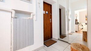 Un couloir avec une porte en bois, un radiateur blanc et un miroir qui reflète une chambre à coucher avec un lit et un bureau.