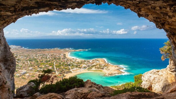 Blick auf eine Küstenstadt durch eine Felsöffnung, mit freiem Blick auf das Meer und einen Sandstrand.