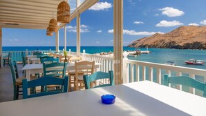 Ein Restaurant am Meer mit Außenbereich, weißen Tischen und blauen Stühlen, mit Blick auf das klare blaue Meer und einen felsigen Hügel.