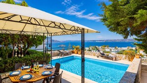 A poolside dining area with a table set for two, a covered seating area, and a view of the sea. - Villa Ikaros in Plaka (Apokoronas)