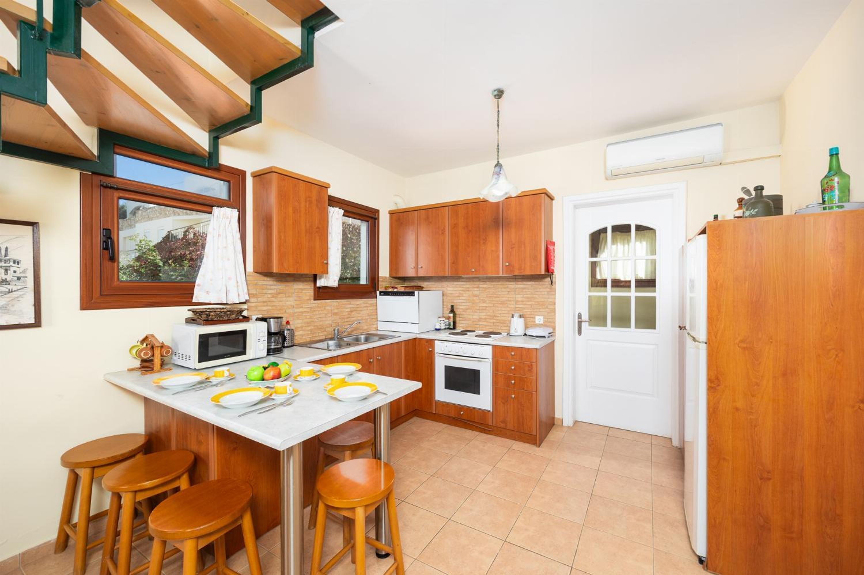 A compact kitchen with wooden cabinets, a white countertop, a built-in oven, a microwave, and a sink. There are three stools at the counter.