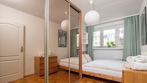 Un dormitorio con piso de madera, cama con ropa de cama blanca, mesita de noche con lámpara, espejo y ventana con cortinas.