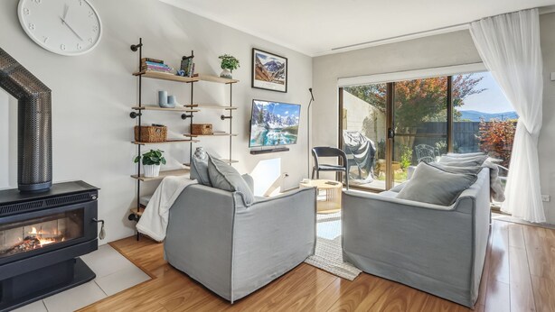 A modern living room with a fireplace, grey sofas, a TV, and a large window with a view of trees and mountains. - Albi Celeste - 2/9 Park Road (Jindabyne)
