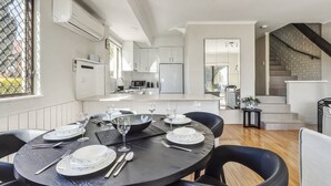 A modern kitchen with white cabinets, a central island, and a dining area with a round table set for six. - Albi Celeste - 2/9 Park Road (Jindabyne)