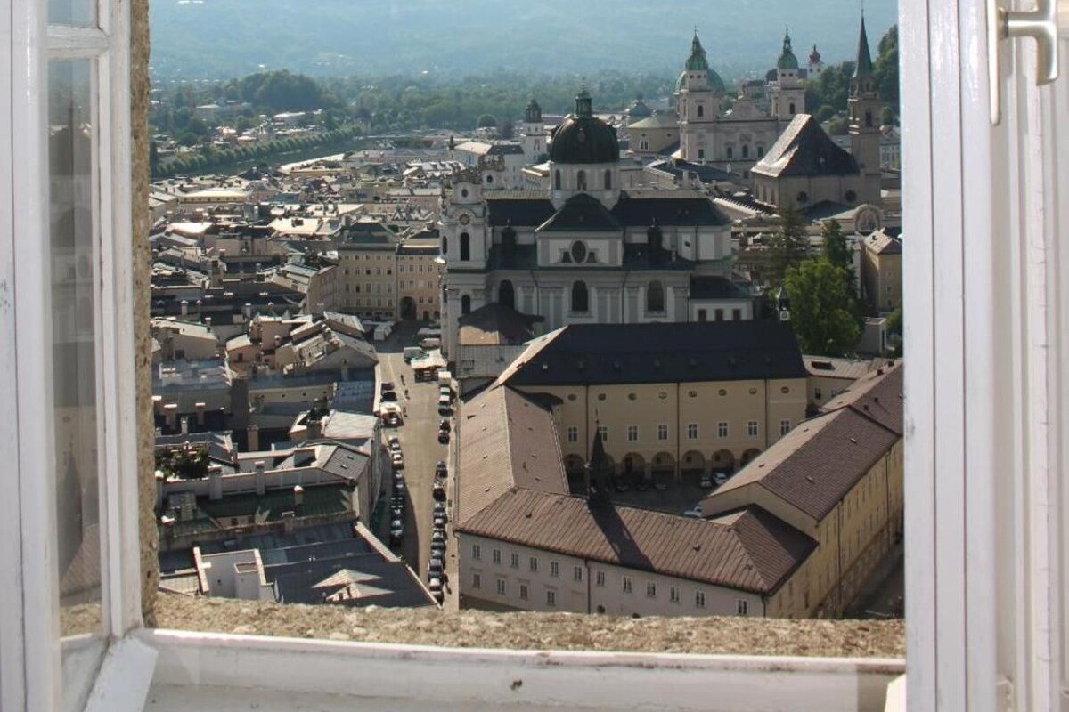 Blick durch ein Fenster auf eine Stadtlandschaft mit historischer Architektur und Bergen im Hintergrund.