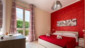 A hotel room with a bed, a bedside table, a window with curtains, a red accent wall, and a hanging light fixture. - Villa Tiffany with Private Terrace, Wi-Fi, Air Conditioning, and Garden View (Campofelice di Roccella)