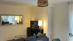 Un salon comprenant un canapé d’angle, une table à manger avec des chaises, un téléviseur posé sur un meuble et un luminaire suspendu.